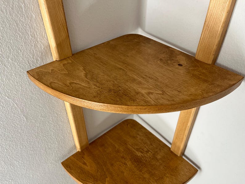 May include: A two-tiered corner shelf made of light-colored wood. The shelves are triangular with rounded edges, supported by vertical and angled wooden supports. The wood grain is visible, and the shelf is mounted on a white wall.