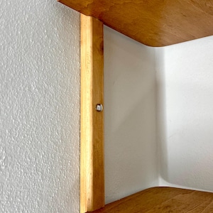 May include: A wooden corner shelf with two curved shelves and a vertical support. The shelves and support are made of light-colored wood, and the shelf is mounted on a white wall. A small metal fastener is visible on the support.
