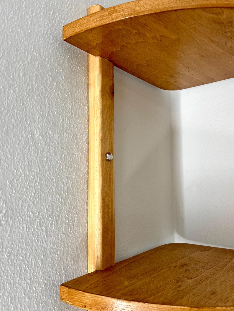 May include: A wooden corner shelf with two curved shelves and a vertical support. The shelves and support are made of light-colored wood, and the shelf is mounted on a white wall. A small metal fastener is visible on the support.