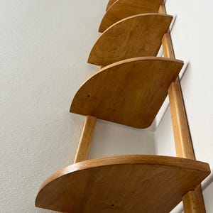 May include: A wooden wall shelf with five semi-circular shelves. The shelves are made of light-colored wood and are supported by vertical wooden supports. The shelf is mounted on a white wall, creating a minimalist and functional design.