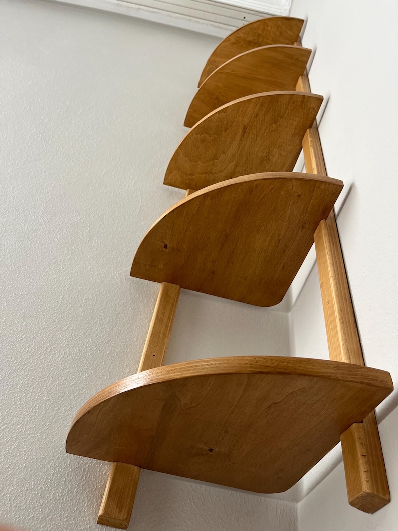 May include: A wooden wall shelf with five semi-circular shelves. The shelves are made of light-colored wood and are supported by vertical wooden supports. The shelf is mounted on a white wall, creating a minimalist and functional design.