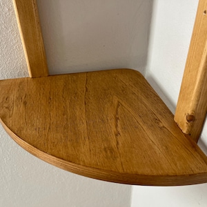 May include: A wooden corner shelf with a semi-circular front edge. The shelf is made of light brown wood and is supported by two vertical wooden supports. The shelf is mounted on a white wall.