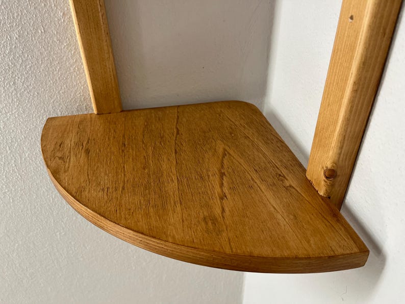 May include: A wooden corner shelf with a semi-circular front edge. The shelf is made of light brown wood and is supported by two vertical wooden supports. The shelf is mounted on a white wall.