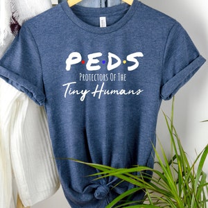 May include: A blue t-shirt with white text that reads "P.E.D.S. Protectors of the Tiny Humans". The text is in a stylized font reminiscent of the popular television show "Friends".