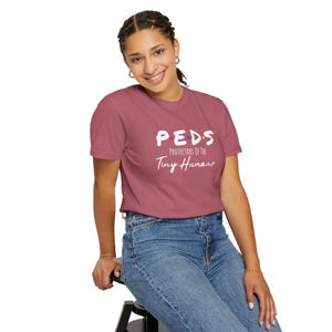May include: A dusty rose t-shirt with the text "PEDS Protectors of the Tiny Humans" in white. The model is wearing blue jeans and is seated on a black stool. The shirt is short-sleeved.