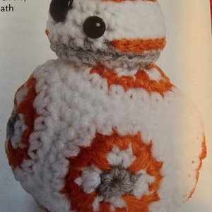 May include: A handmade crochet BB-8 droid. The droid is primarily white with orange and gray accents. It features large black eyes and a detailed, textured design, perfect for Star Wars fans.