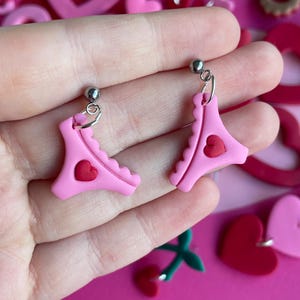 May include: Pink polymer clay earrings shaped like underwear with a red heart detail. The earrings are on silver hoops. Other Valentine's Day themed polymer clay jewelry is in the background, including hearts and cookies.