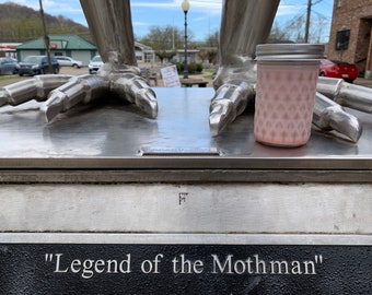 May include: A close-up of a silver statue of a creature with large feet. The statue is labelled "Legend of the Mothman". A pink jar with a silver lid is sitting on the statue's base.