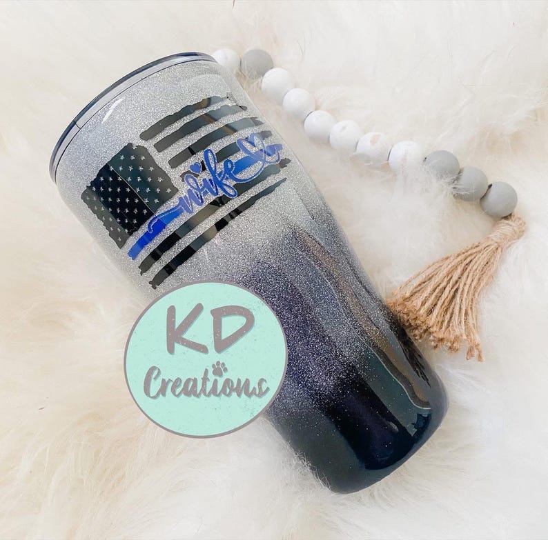 May include: A glitter-covered tumbler with a design featuring a distressed American flag with a blue line and the word "wife" in blue script. The tumbler transitions from silver to dark blue.