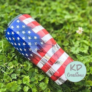May include: A stainless steel tumbler featuring a patriotic design of the American flag. The tumbler has a blue section with white stars and red and white stripes. The tumbler is lying on a bed of green grass.