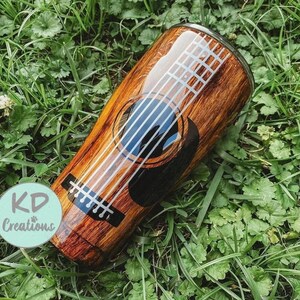 May include: A stainless steel tumbler with a wood grain design and a guitar graphic. The guitar features white strings, a black sound hole, and a blue interior. The tumbler is cylindrical and has a glossy finish. The text "KD Creations" is on the side.