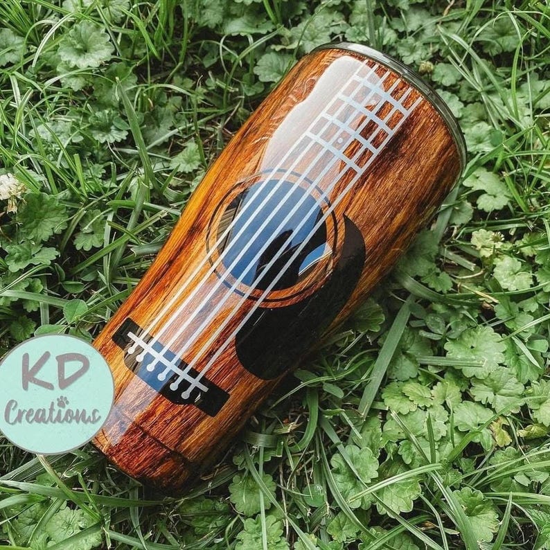 May include: A stainless steel tumbler with a wood grain design and a guitar graphic. The guitar features white strings, a black sound hole, and a blue interior. The tumbler is cylindrical and has a glossy finish. The text "KD Creations" is on the side.