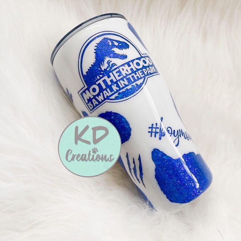 May include: White tumbler with a blue dinosaur logo that reads "Motherhood is a walk in the park." The tumbler has blue glitter accents and the hashtag #oym. A teal circle with "KD Creations" is also visible.