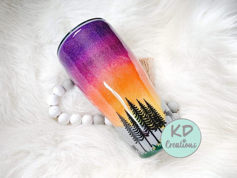 May include: A glittery tumbler with a sunset design. The top of the tumbler is purple, transitioning to orange and yellow. Black trees are silhouetted against the sunset. The tumbler has the text "KD Creations" on it.