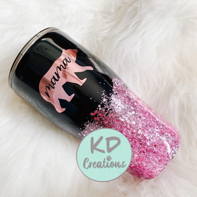 May include: A black tumbler with a pink glitter bottom and a rose gold bear graphic that says "mama". The tumbler has a stainless steel rim and a black lid. The tumbler is approximately 10 inches tall.