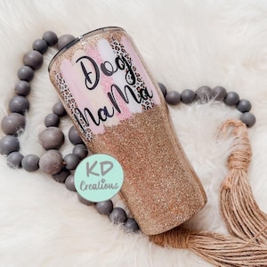 May include: A glitter-covered tumbler with the words "Dog Mama" in black script. The tumbler features pink and white brushstrokes, leopard print accents, and a gold glitter base. A light green circle with the text "KD Creations" is also visible.