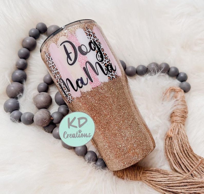 May include: A glitter-covered tumbler with the words "Dog Mama" in black script. The tumbler features pink and white brushstrokes, leopard print accents, and a gold glitter base. A light green circle with the text "KD Creations" is also visible.