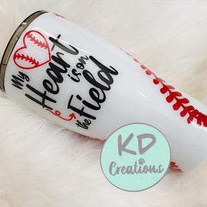May include: White tumbler with a baseball theme. The tumbler features a red heart with baseball stitching, the phrase "My Heart is on the Field," and red baseball stitching details. A teal circle with "KD Creations" is also visible.