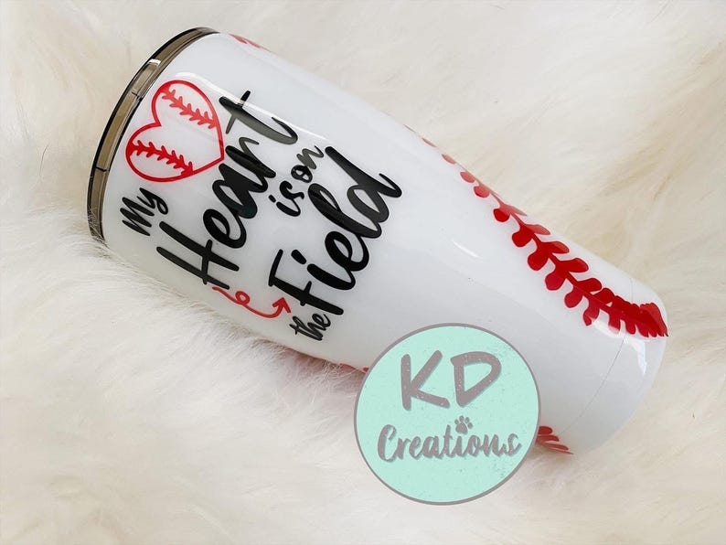 May include: White tumbler with a baseball theme. The tumbler features a red heart with baseball stitching, the phrase "My Heart is on the Field," and red baseball stitching details. A teal circle with "KD Creations" is also visible.