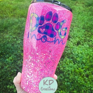 May include: A pink glitter tumbler with a black lid. The tumbler features the text "Dog Mom" with a paw print design in blue, yellow, and plaid patterns. A round sticker with the text "KD Creations" is visible.