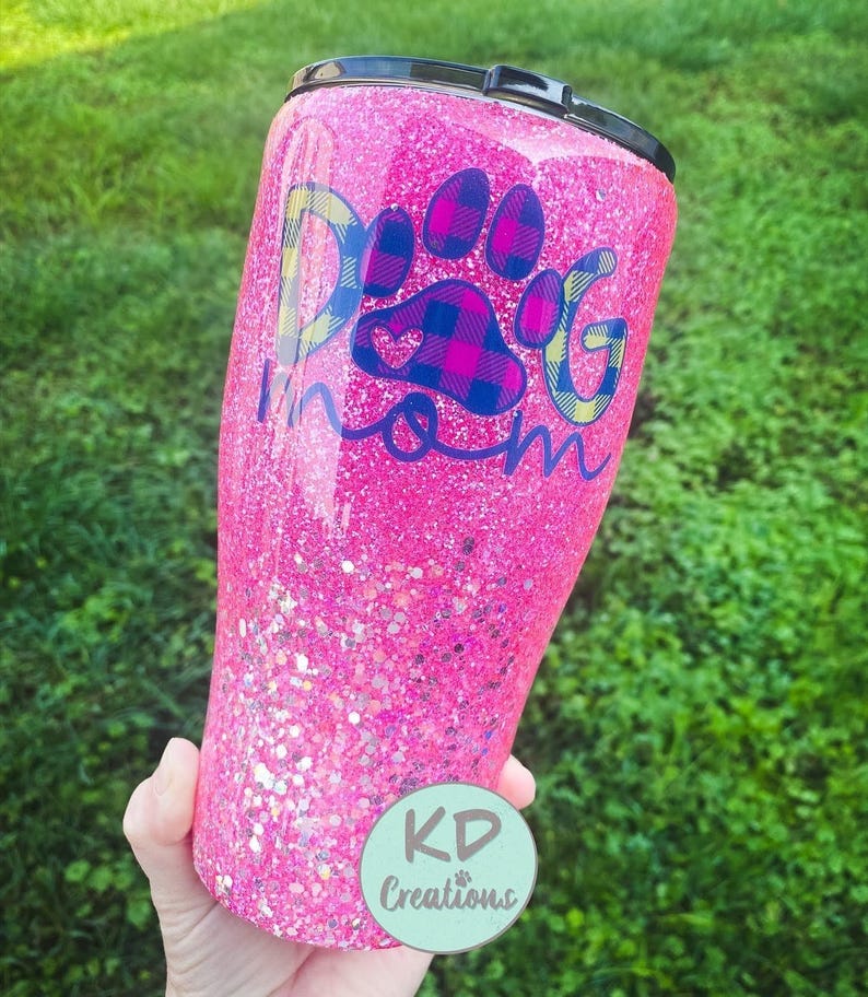 May include: A pink glitter tumbler with a black lid. The tumbler features the text "Dog Mom" with a paw print design in blue, yellow, and plaid patterns. A round sticker with the text "KD Creations" is visible.