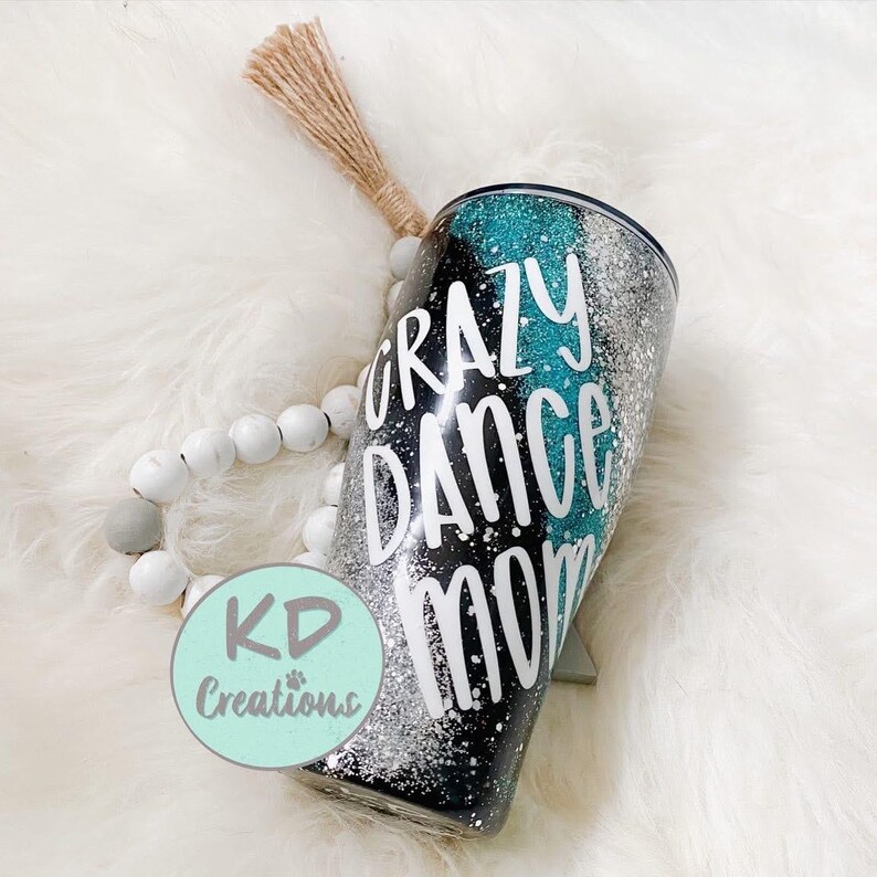 May include: A glitter-covered tumbler with the words "Crazy Dance Mom" in white. The tumbler has a black base with silver and teal accents. A light brown tassel and a white beaded bracelet are also visible.