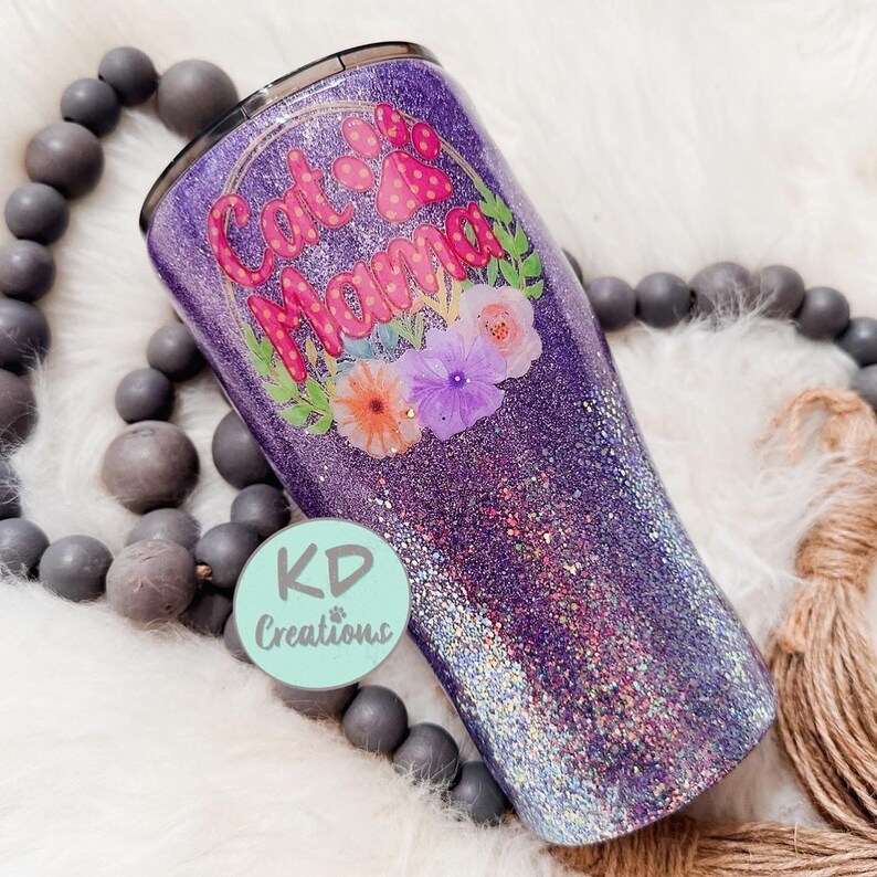 May include: A purple glitter tumbler with the words "Cat Mama" in pink, surrounded by a floral design. The tumbler has a black lid and a light blue logo that says "KD Creations".