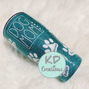 May include: Teal glitter tumbler with the words "DOG MOM" in white. White paw prints and dog names are featured on the tumbler. A round sticker with the text "KD Creations" is on the bottom of the tumbler.