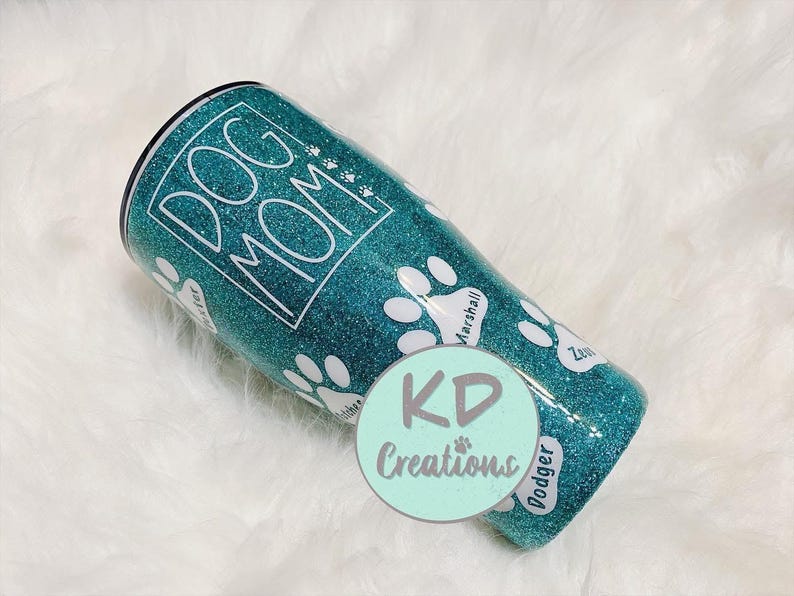 May include: Teal glitter tumbler with the words "DOG MOM" in white. White paw prints and dog names are featured on the tumbler. A round sticker with the text "KD Creations" is on the bottom of the tumbler.