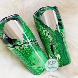 May include: Two stainless steel travel tumblers with a green and black marbled design. Each tumbler has a silver lid and a silver cutout design. The tumblers are on a white, fluffy surface. The logo "KD Creations" is visible.