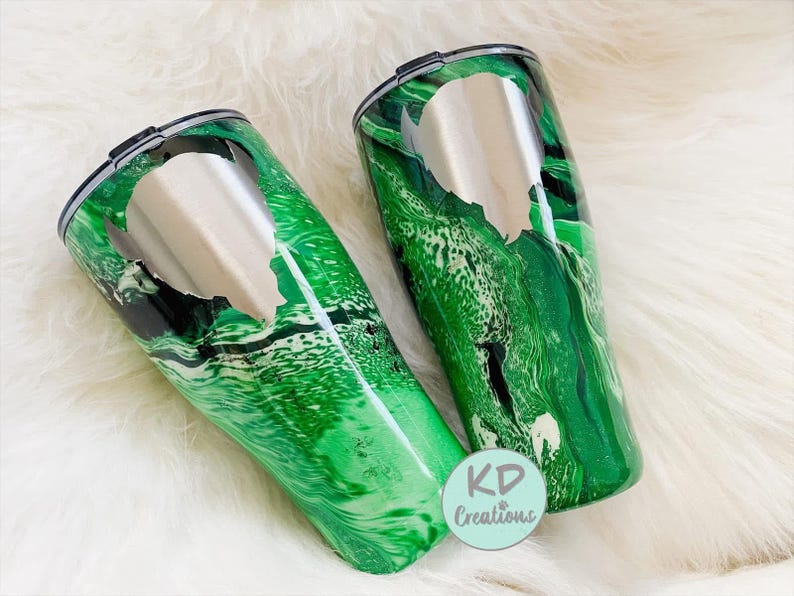 May include: Two stainless steel travel tumblers with a green and black marbled design. Each tumbler has a silver lid and a silver cutout design. The tumblers are on a white, fluffy surface. The logo "KD Creations" is visible.