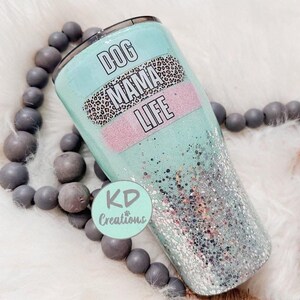 May include: A teal tumbler with glitter and the words "DOG MAMA LIFE" in a black font. The tumbler has a leopard print band and a pink band. The tumbler is surrounded by gray beads.