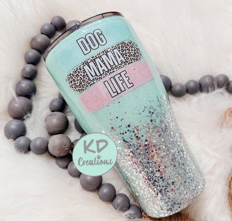 May include: A teal tumbler with glitter and the words "DOG MAMA LIFE" in a black font. The tumbler has a leopard print band and a pink band. The tumbler is surrounded by gray beads.