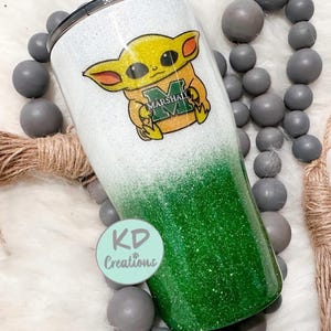 May include: A glitter-covered tumbler with a white and green ombre design. The tumbler features a cartoon character with the text "MARSHALL" on its chest. The tumbler also has a black lid and the text "KD Creations" on a round sticker.