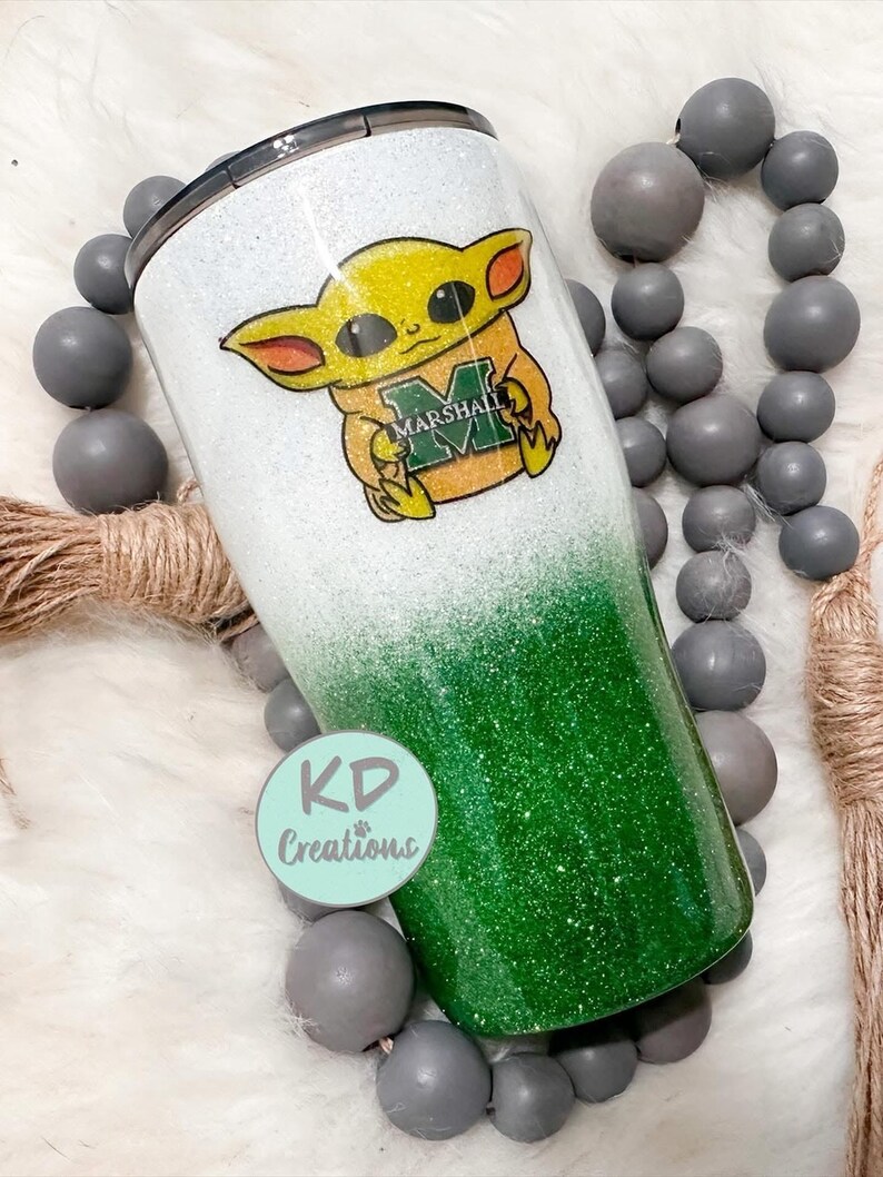 May include: A glitter-covered tumbler with a white and green ombre design. The tumbler features a cartoon character with the text "MARSHALL" on its chest. The tumbler also has a black lid and the text "KD Creations" on a round sticker.