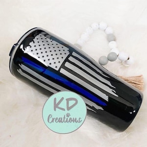 May include: Black tumbler with a distressed American flag design, featuring a blue line. The tumbler has a black lid and a silver and white flag design. The text "KD Creations" is on the bottom.
