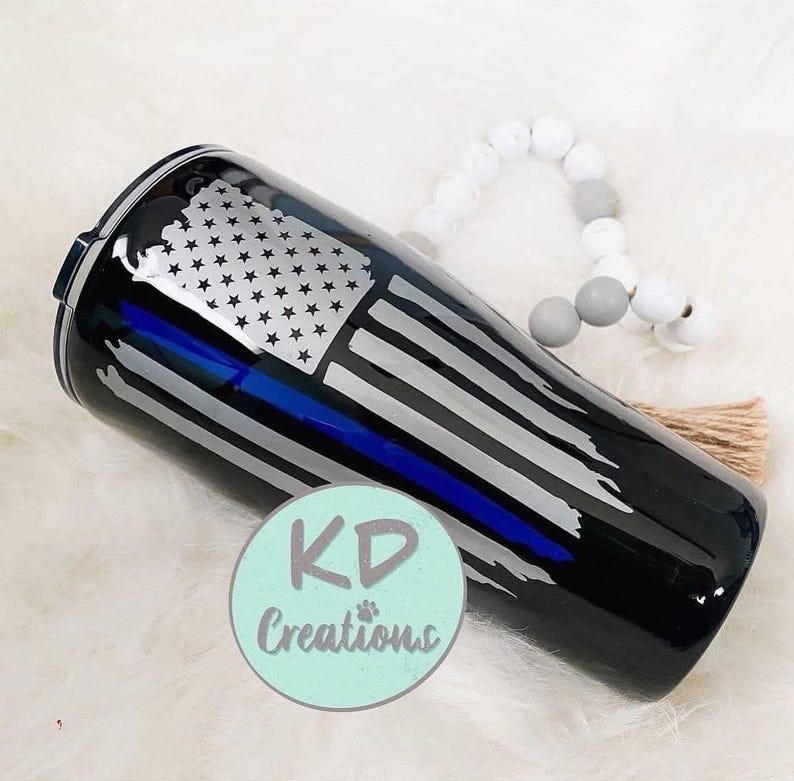 May include: Black tumbler with a distressed American flag design, featuring a blue line. The tumbler has a black lid and a silver and white flag design. The text "KD Creations" is on the bottom.