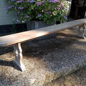 May include: A long, narrow wooden bench with a light brown, weathered finish. The bench features a smooth, rounded-edge seat supported by two vertical legs, each designed with a decorative cutout shape resembling a stylised clover or cross.