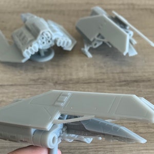 May include: Three gray miniature spaceship models are displayed. The models have detailed designs, including wings, engines, and other mechanical features. The spaceships are likely 3D-printed and are on a wooden surface.