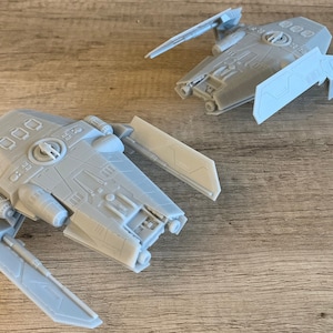 Peut inclure: Deux mod&egrave;les de vaisseaux spatiaux imprim&eacute;s en 3D gris avec des designs d&eacute;taill&eacute;s et des ailes. Les mod&egrave;les sont sur une surface en bois.