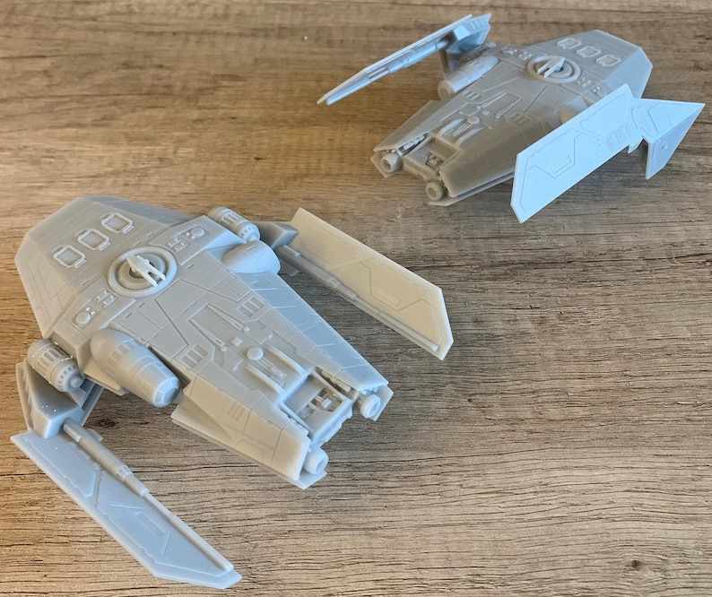 Peut inclure: Deux mod&egrave;les de vaisseaux spatiaux imprim&eacute;s en 3D gris avec des designs d&eacute;taill&eacute;s et des ailes. Les mod&egrave;les sont sur une surface en bois.
