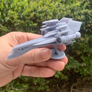 Pode incluir: Um modelo de nave espacial futurista impresso em 3D de cor cinza com v&aacute;rios motores e um nariz longo e pontudo. A nave espacial est&aacute; montada em uma base circular.