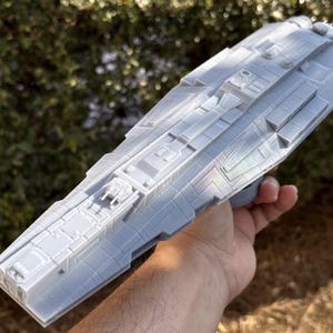 Peut inclure: Maquette de vaisseau spatial gris clair avec des détails de surface complexes, tenue dans une main. Le modèle présente un corps rectangulaire avec des panneaux surélevés, des tourelles et des composants de moteur. Le design suggère un thème de science-fiction.