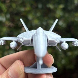 Puede incluir: Maqueta de avi&oacute;n de combate gris claro con alas detalladas, aletas de cola y accesorios para misiles. El modelo est&aacute; montado sobre una peque&ntilde;a base circular. El fondo es follaje verde borroso.