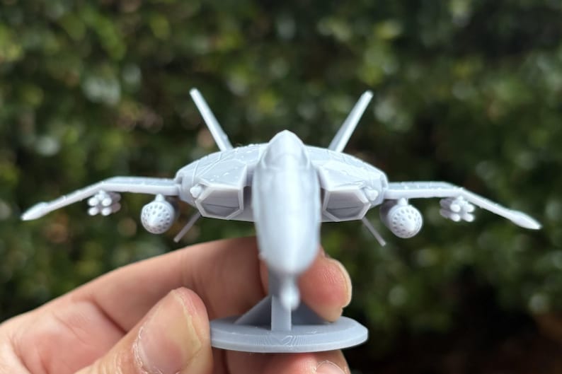 Puede incluir: Maqueta de avi&oacute;n de combate gris claro con alas detalladas, aletas de cola y accesorios para misiles. El modelo est&aacute; montado sobre una peque&ntilde;a base circular. El fondo es follaje verde borroso.