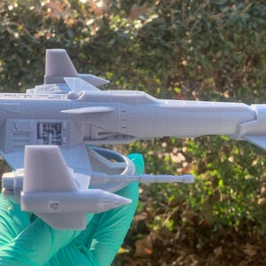 May include: A gray 3D printed model of a spaceship with a detailed design. The model has a long, narrow body with a pointed nose and two wings. The wings are angled upwards and have a slight curve to them. The model is sitting on a green surface.