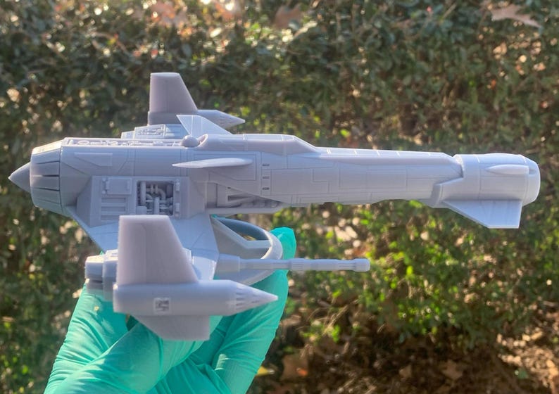 May include: A gray 3D printed model of a spaceship with a detailed design. The model has a long, narrow body with a pointed nose and two wings. The wings are angled upwards and have a slight curve to them. The model is sitting on a green surface.