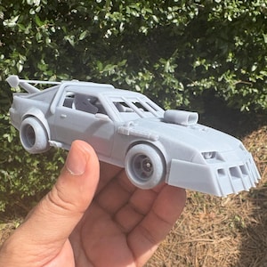 May include: A gray, 3D-printed model car with a spoiler, wide tires, and a hood scoop. The car has a futuristic design with a low front end and a roll cage. The model is held in a hand against a background of green foliage and brown grass.