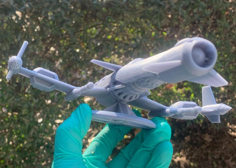 May include: A gray 3D printed model of a futuristic spaceship with a large engine and a small wing on the side. The spaceship is sitting on a circular base.