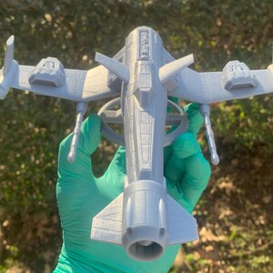May include: A gray 3D printed model of a futuristic spaceship with a long, cylindrical body and two wings extending out to the sides. The spaceship has a detailed design with a variety of panels and hatches.